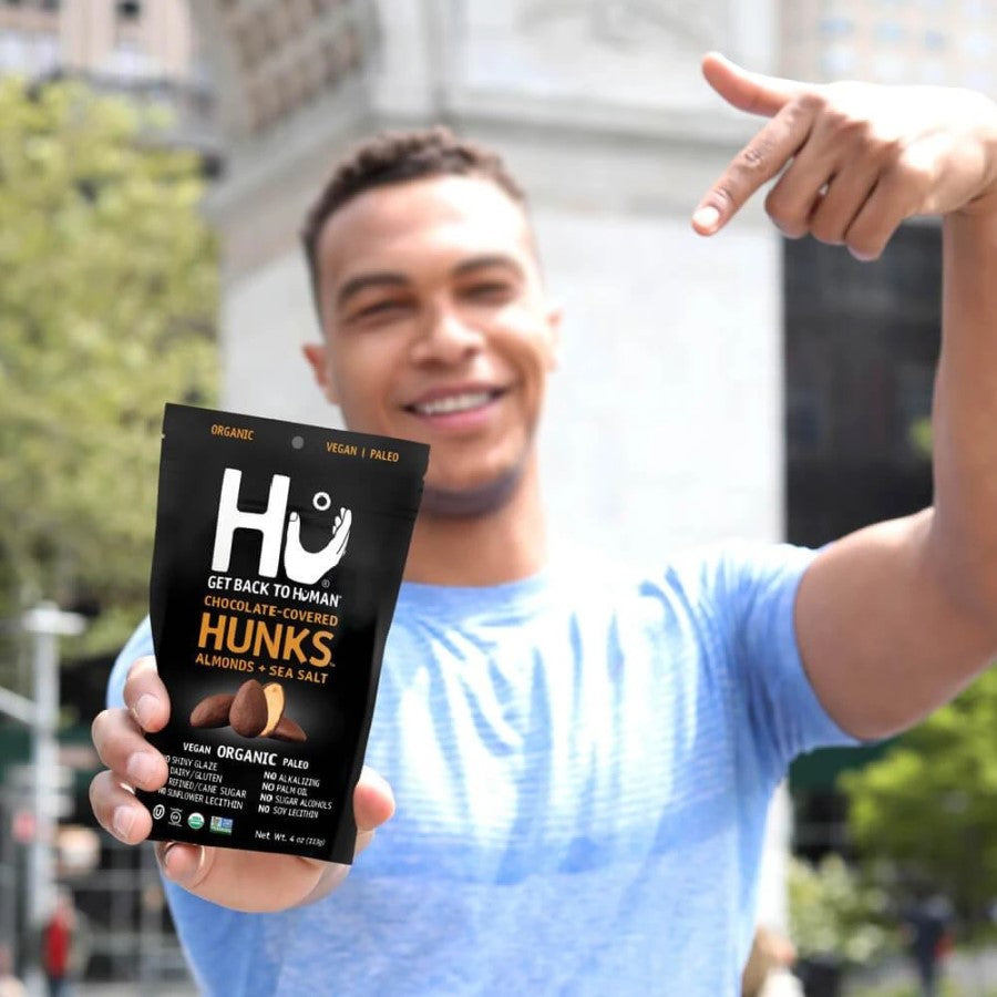 Guy Holding Up And Pointing To Bag Of Hu Hunks Organic Chocolate Covered Almonds