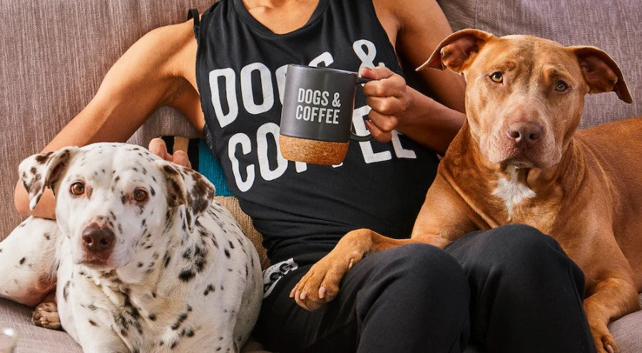 Woman Wearing Grounds And Hounds Apparel Drinking Out Of Dogs & Coffee Mug Sitting On Sofa With Two Dogs