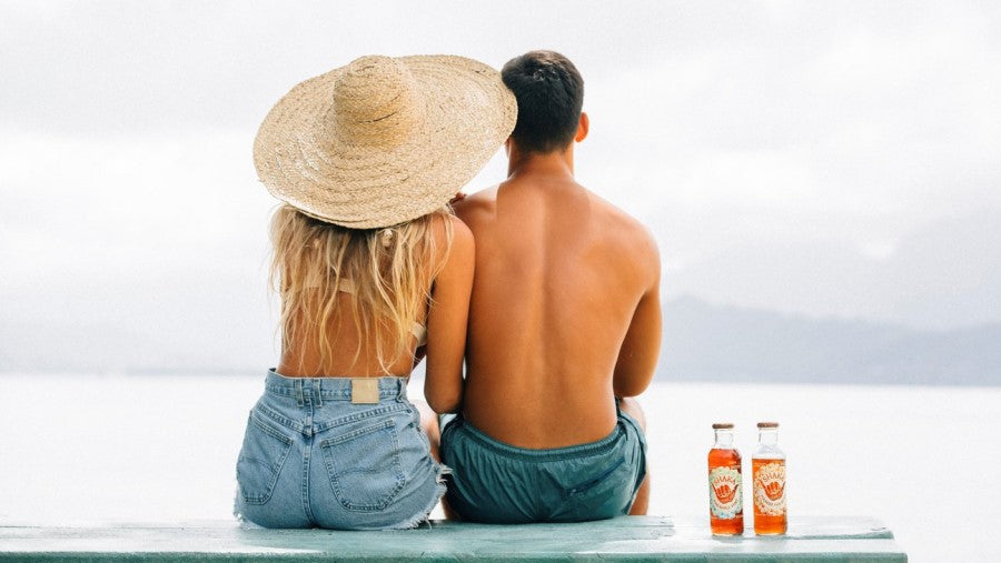 Couple Sitting On A Picnic Table At The Beach In Hawaii With Two Bottles Of Shaka Tea Made With Mamaki Leaves For Healthy Hydration Zero Caffeine