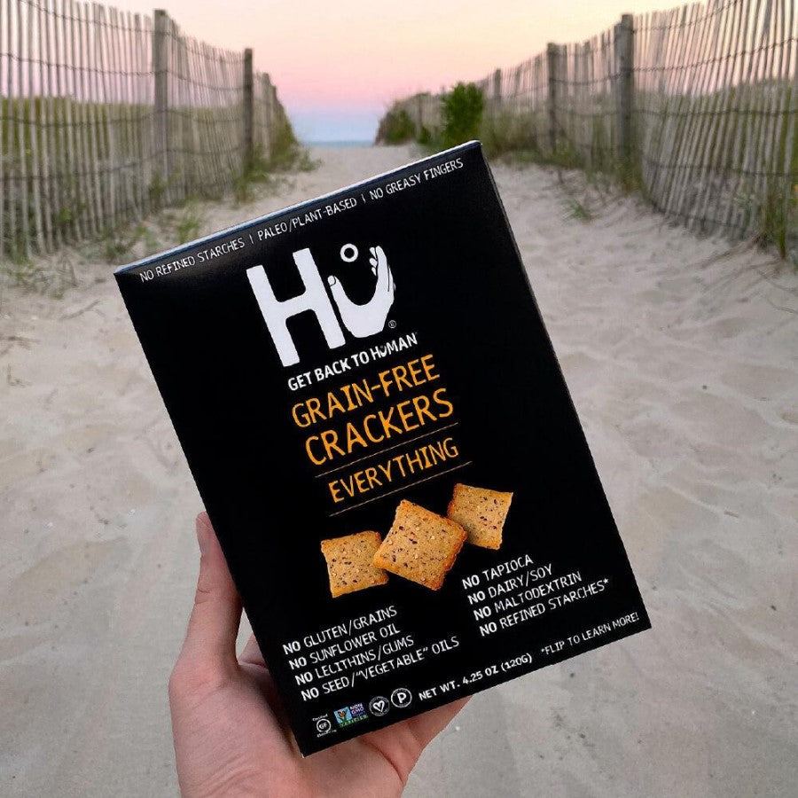 Hand Holding A Box Of Hu Everything Grain Free Crackers On The Beach At Sunset
