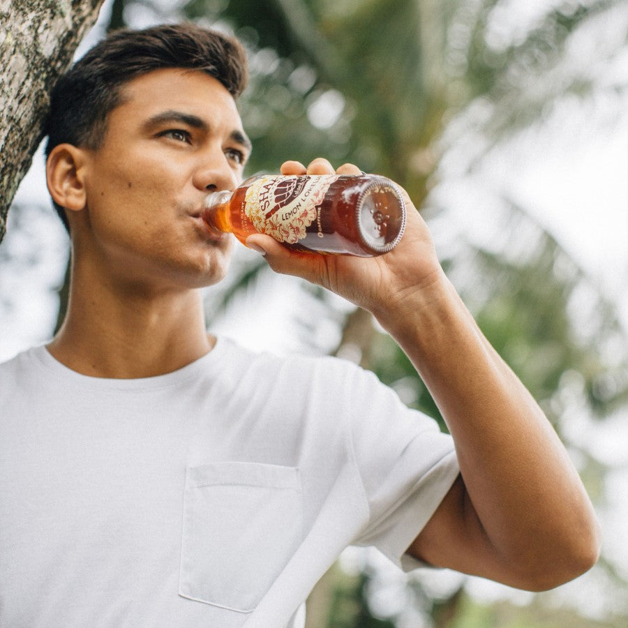 Man Drinking A Bottle Of Shaka Lemon Lokelani Rose Tea Outdoors By Palm Trees In Hawaii