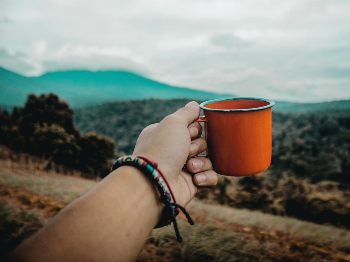 Hand Holding Orange Metal Camp Cup Of Organic Coffee Outdoors