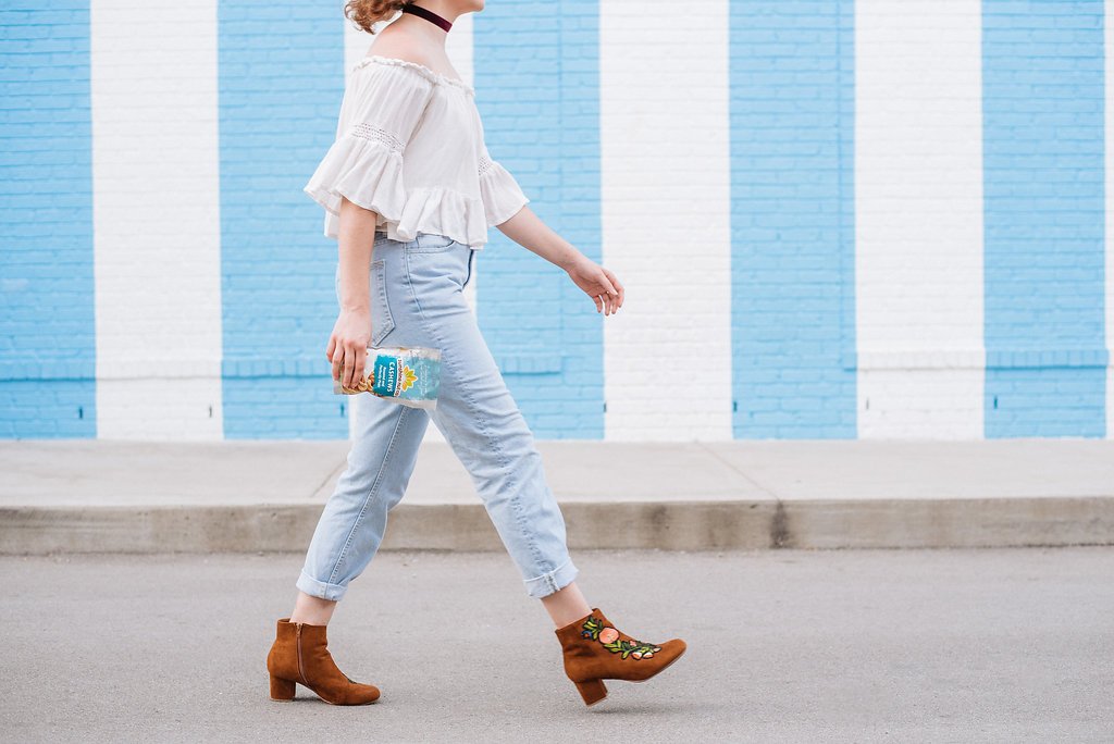 Woman Walking In Front Of Blue And White Stripe Wall Holding Bag Of Perfectly Plain Roasted Cashews From Sunshine Nut