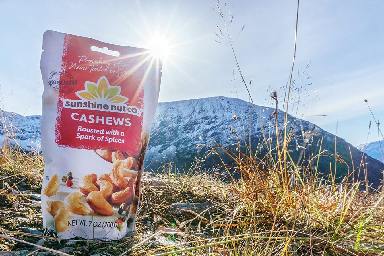 7 Ounce Sunshine Nut Co. Bag Of Cashews Roasted With A Spark Of Spices Sitting In Grass Near Mountains Outdoors In Sunshine