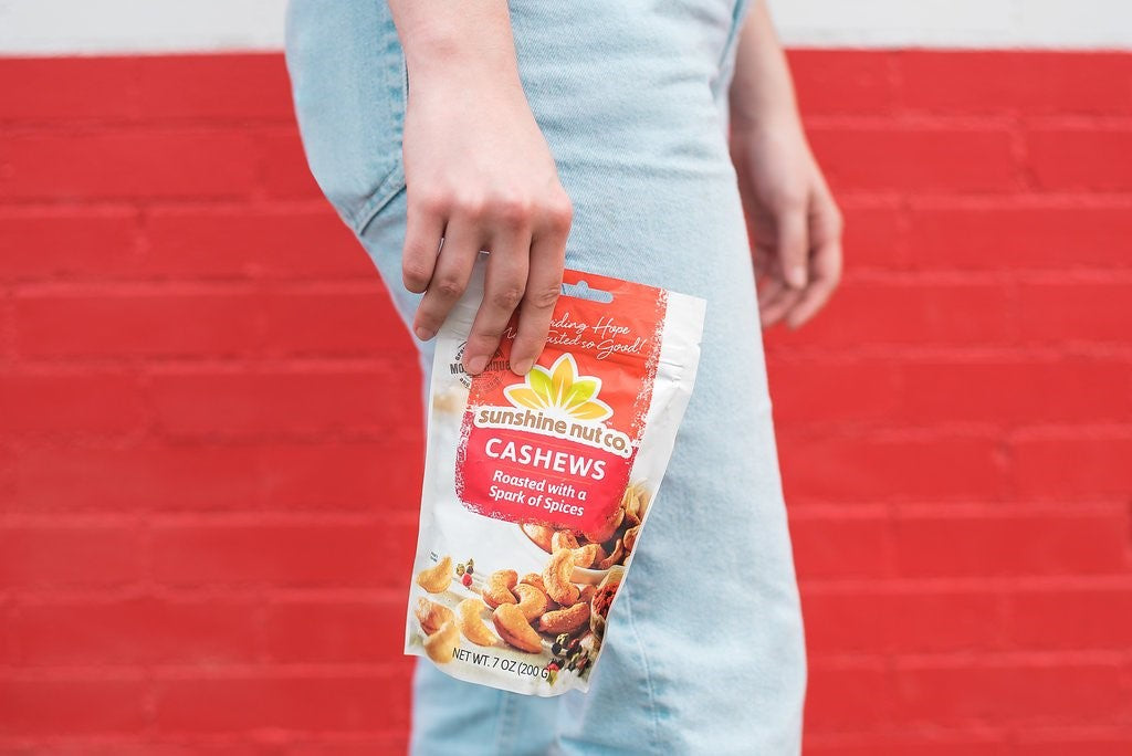 Woman's Hand Holding Seven Ounce Bag Of Spicy Cashews From Sunshine Nut Company Roasted With Spark Of Spices In Front Of Red Brick Wall