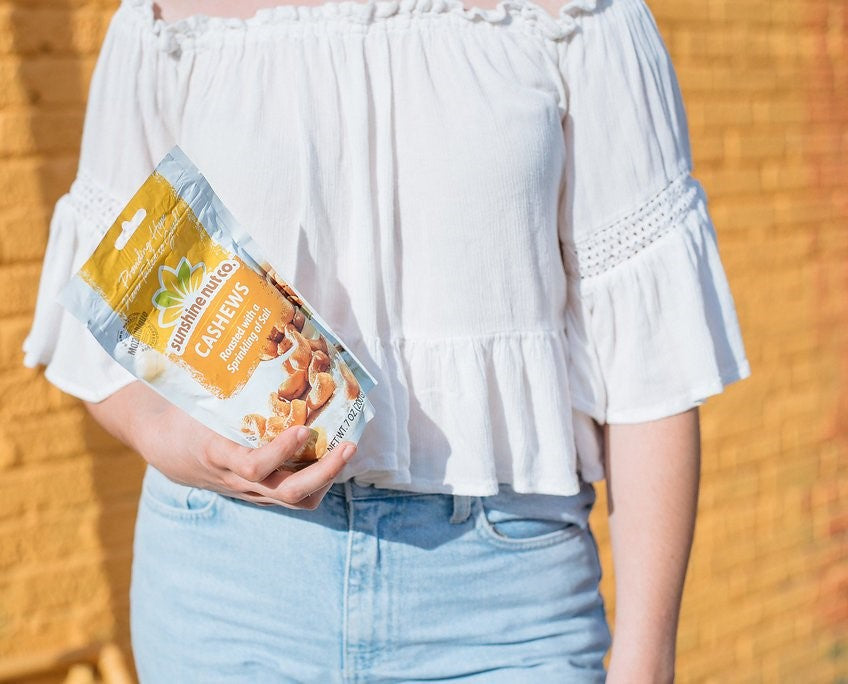Woman Outdoors Near Yellow Sunny Wall Holding Bag Of Sunshine Nut Co Cashews With Sprinkling Of Salt