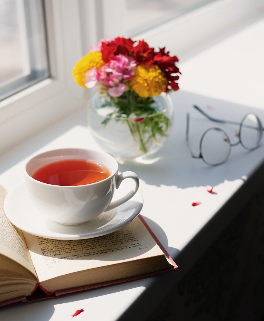 Beautiful Cup Of Tea In Sunlight With Book Glasses And Vase Of Colorful Flowers