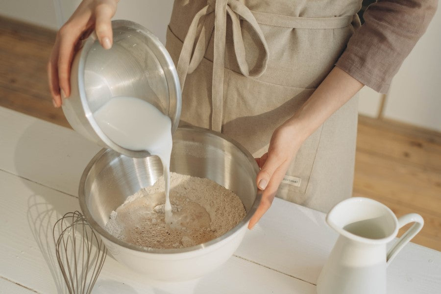 Woman In Kitchen Wearing Apron Pouring Dairy Free Plant Based Milk Into Recipe Unsweetened Better Than Milk Calcium Rice Drink From Terra Powders