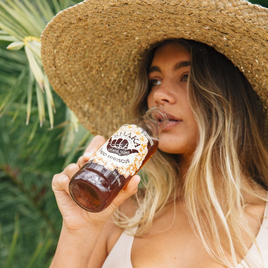 Woman Outdoors In Summer Hat Drinking A Bottle Of Shaka Iced Tea Mango Hibiscus Flavor