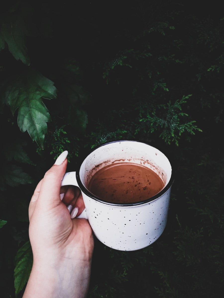 Woman's Hand With White Nails Holding White Metal Camp Mug Of Coffee With Cocoa Outdoors In Front Of Greenery