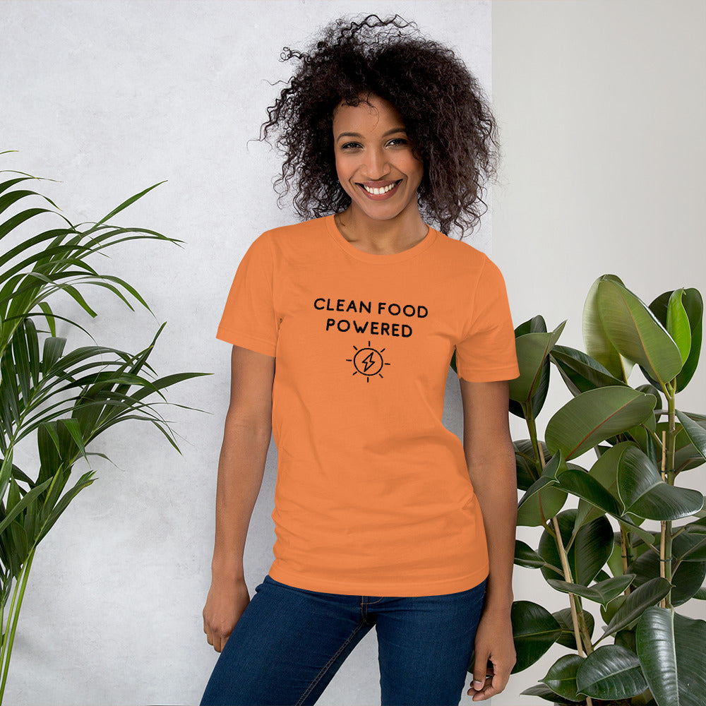 Happy Woman Standing By Plants Wearing Clean Food Powered Tee Shirt In Burnt Orange From Terra Powders